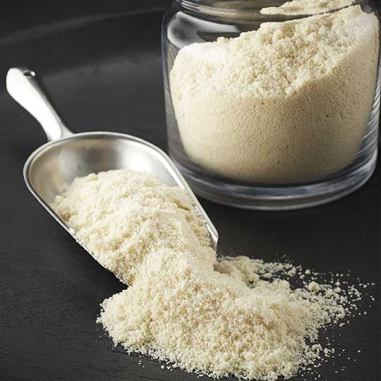 Stainless steel scoop with finely milled almond flour spilling out onto black table with clear jar full of almond flour beside scoop