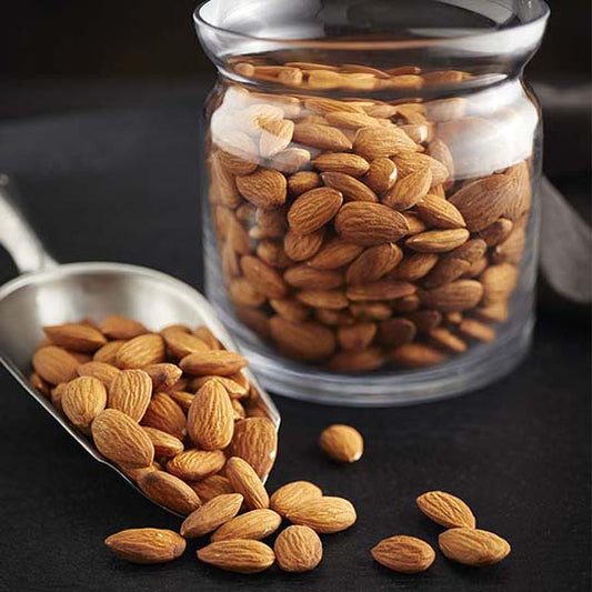 golden brown whole almonds spilling out of stainless steel scoop with whole almonds filling a clear glass jar