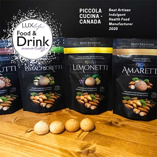 One bag of each flavour of piccola cucina italian almond amaretti with each of the cookies on wood table in front of bags at lux food and drink awards