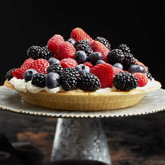 Almond pie crust full of cream cheese base and topped with heaping amounts of blueberries blackberries raspberries and strawberries sitting on silver dessert plate with black background