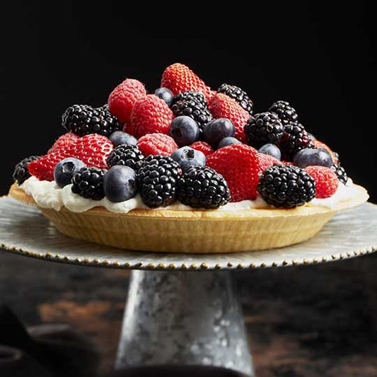Almond pie crust full of cream cheese base and topped with heaping amounts of blueberries blackberries raspberries and strawberries sitting on silver dessert plate with black background