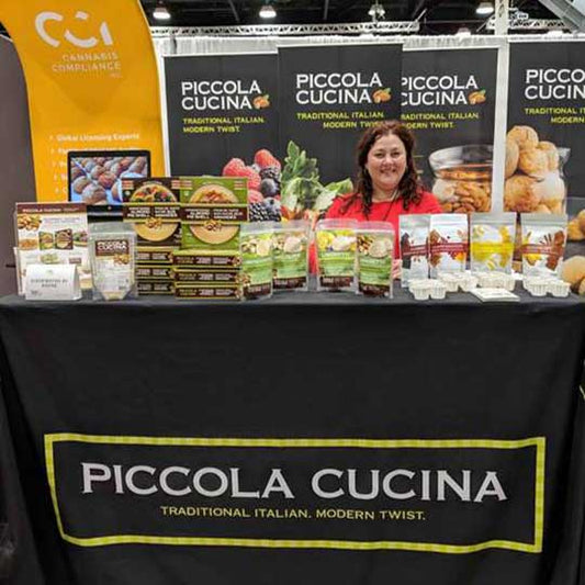 Owner Pina Romolo behind Piccola Cucina booth with various product selections on table at Canadian Health Food Assosiation West 