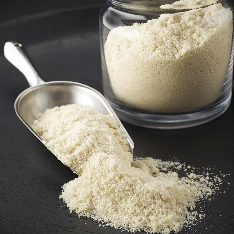 silver scoop with white almond flour spilling out onto black table with clear jar filled with almond flour to the right
